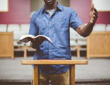 A closeup shot of a male reading the bible near a wooden stand with a blurred background